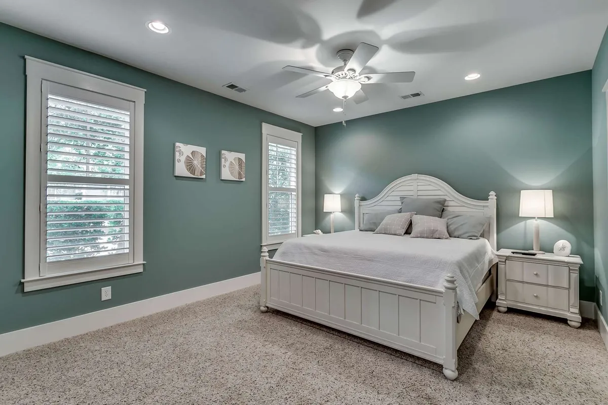 A serene bedroom featuring a crisp white bed against calming green walls, with a bedside table topped by a lamp and a window that lets in gentle natural light. The setting emphasizes a peaceful atmosphere ideal for reading, relaxing, or a restful night's sleep.