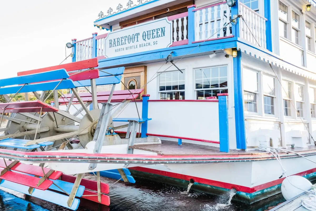 A brightly colored paddlewheel riverboat docked by the water, featuring playful signage and vibrant architecture. The deck looks inviting for group dining, live music, and sunset sightseeing, evoking a sense of local adventure and group outings.