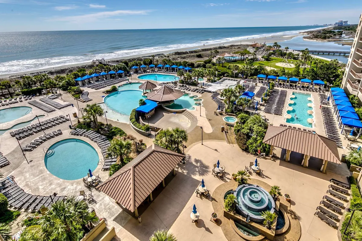 A sprawling resort pool complex with beachfront views, palm trees, loungers, and umbrellas. The tranquil, vacation-like atmosphere invites swimming, sunbathing, and poolside socializing, suggesting seamless access from home to resort amenities.