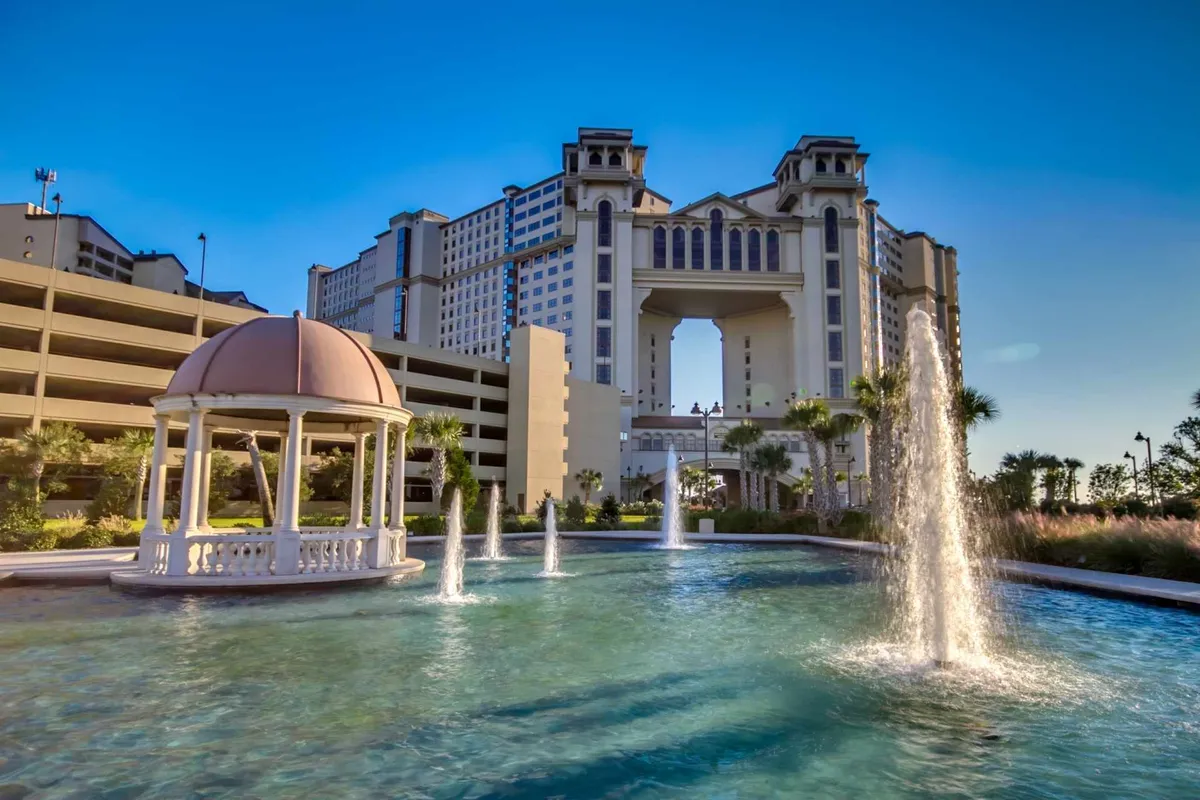 This exterior scene showcases a tranquil setting with a fountain, gazebo, and inviting building archway. The sun sets in the background, casting warm light over the water and lush landscaping, inviting relaxation and social interaction by the water’s edge.