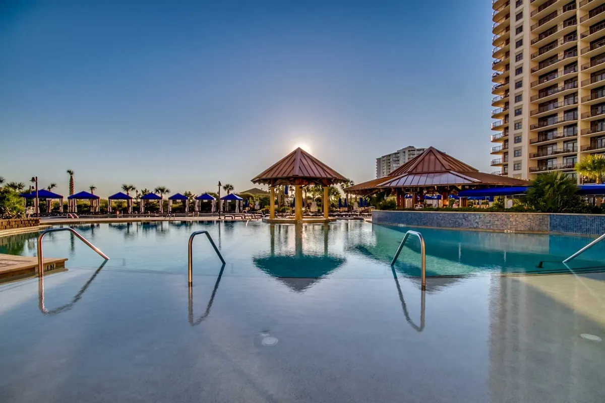 This image showcases an inviting swimming pool surrounded by thatched-roof cabanas, palm trees, and comfortable lounge chairs with a beautiful sunset in the background. The scene is tranquil yet lively, conveying a resort-style atmosphere perfect for families seeking relaxation and fun moments together.