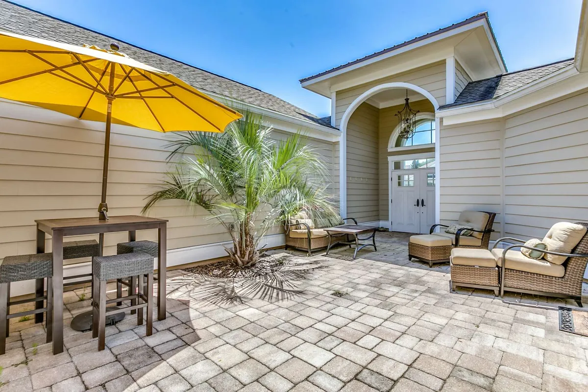 A cheerful and inviting courtyard scene featuring a yellow umbrella, palm tree, comfortable outdoor seating, and the entrance door. The area is bathed in sunlight, suggesting a perfect spot for relaxing with coffee in the morning or hosting casual get-togethers in the evening.