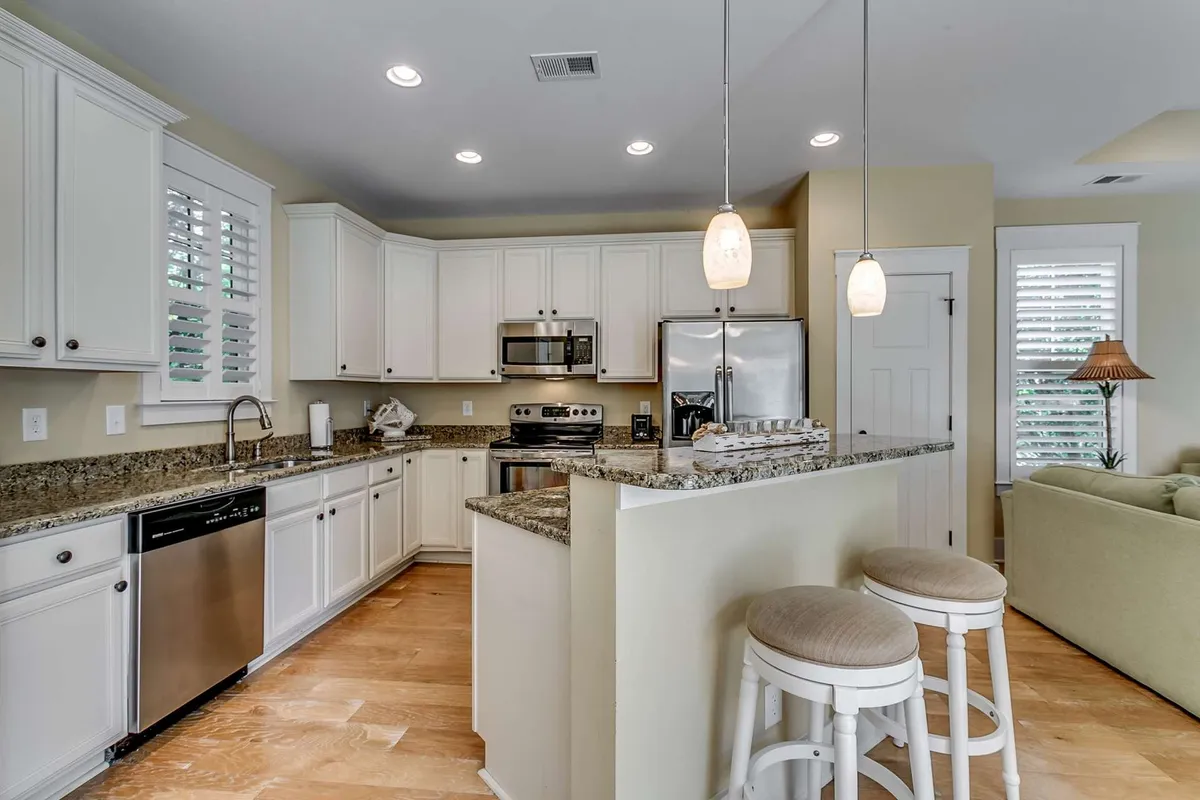 This image features a contemporary kitchen with granite countertops, stainless steel appliances, and an inviting island with seating. The open layout encourages cooking and socializing, highlighted by stylish light fixtures. Family members or guests can easily gather around the island, making it ideal for collaborative meal prep or casual dining.