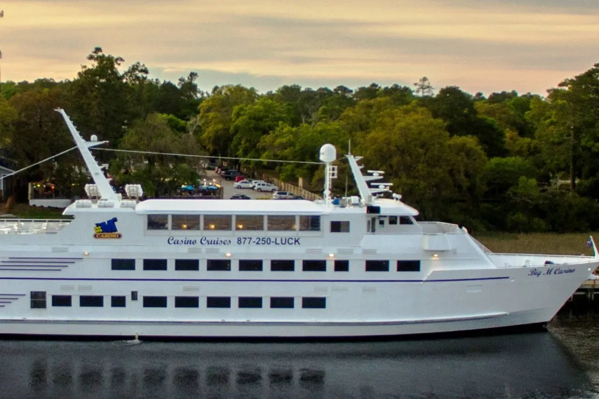 A lively exterior scene featuring a casino cruise ship docked at a marina, surrounded by lush trees. The image captures the ship's decks, inviting guests to enjoy dining, entertainment, and sunset views, all set against the backdrop of the marina and natural scenery.