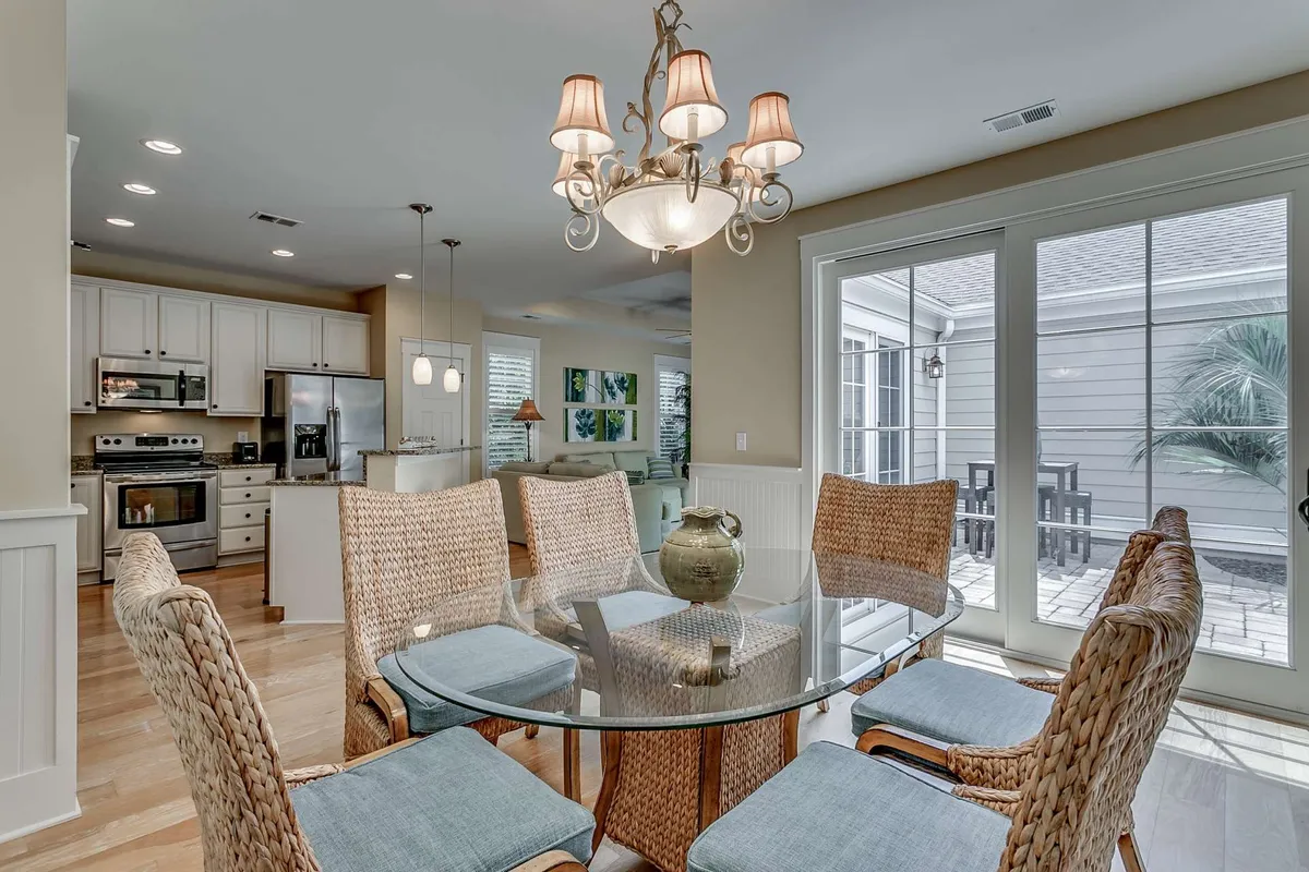 A glass dining table surrounded by woven chairs, illuminated by a stylish chandelier and framed by large windows that fill the room with natural light. The space is clearly set up for group dining or casual meals.