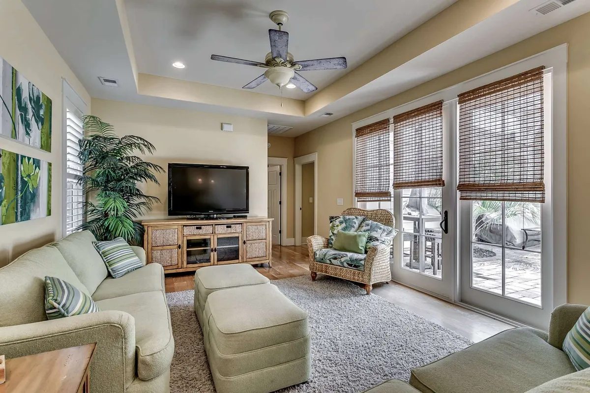 A cozy, open living area with a green sofa, woven chair, television, decorative plant, and large windows allowing natural light to fill the space. The setup encourages relaxation and socializing, with comfortable furnishings and thoughtful decor touches.
