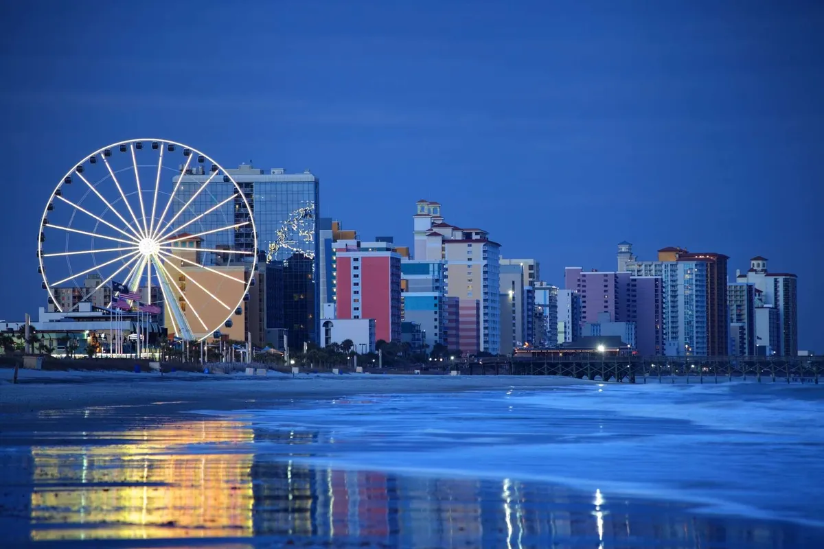 This vibrant exterior image features the iconic Myrtle Beach Ferris wheel, colorful beachfront buildings, and the ocean reflecting the glow of sunset. The lively boardwalk atmosphere is evident, with amusement rides and opportunities for beachside walks.